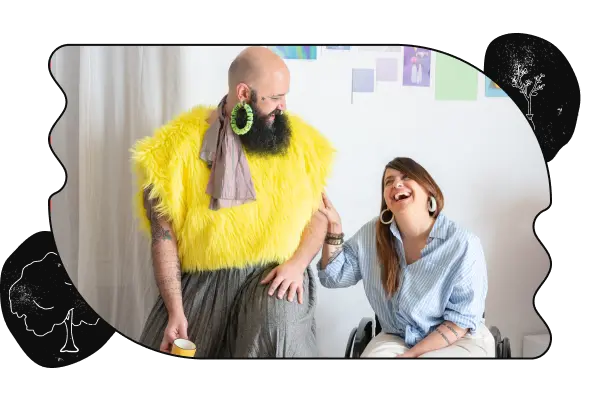 A picture of two people talking and laughing. The person on the left has a beard and is wearing a skirt and fuzzy yellow top. The person on the right is touching their arm while laughing and is sitting in a wheelchair. The image is cropped in a bubbly shape with a black border and tree sketches on the top-left and bottom-right corners.