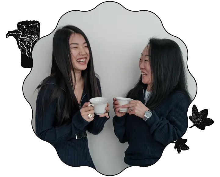 An image of two east-asian women laughing and holding coffee cups. The image is cropped to a wavy-shaped frame with a black border, with a stamp of two mushrooms growing out of a log on the top-left and a stamp of two leaves on the bottom-right.